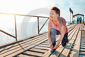 Sport. Happy adult woman in sportswear, tying the laces on her sneakers. Pier and ocean in the background