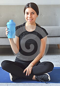 Sport at home concept. Smiling young girl is sitting on mat with sports equipment at home