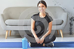 Sport at home concept. Smiling young girl is sitting on mat with sports equipment at home