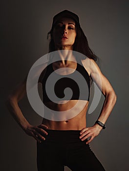 Sport muscular woman posing in black sport bra and cap showing the shoulders, abs and arms, standing on dark shadow studio