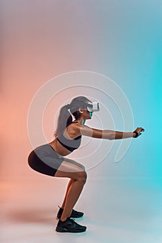 Sport in a new way. Side view of young sporty woman in sports clothing doing squat while wearing virtual reality glasses