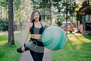 Sport and recration concept. Pleased slim brunette woman wears cropped top and leggings, holds fitball under arm, rolled up