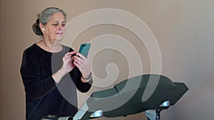 Sport senior woman is training on treadmill in home and looking in mobile phone