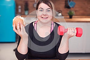 Sport with unhealthy food. combination of active life with fast food. close up photo. fat woman shoeing dumbbell and hamburger
