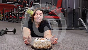 Sport with unhealthy food. Young plus size fat woman puts away barbells and takes a large cake. Young woman looking