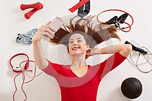 Sport woman smiling at camera of her moderm cell phone, making self picture at gym while laying on floor, young girl being at