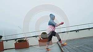 Sport woman in sportswear wears hood doing workout, step cardio exercising training on roof of house