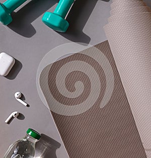 Sport or workout concept flat lay with blue dumbbells, yoga mat and white wireless headphones on the grey background.