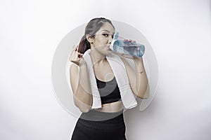 Sportive Asian woman posing with a towel on her shoulder and drinking from a bottle of water, smiling and relaxing after workout