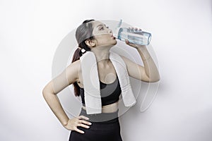 Sportive Asian woman posing with a towel on her shoulder and drinking from a bottle of water, smiling and relaxing after workout