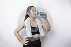 Sportive Asian woman posing with a towel on her shoulder and drinking from a bottle of water, smiling and relaxing after workout