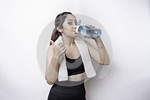 Sportive Asian woman posing with a towel on her shoulder and drinking from a bottle of water, smiling and relaxing after workout