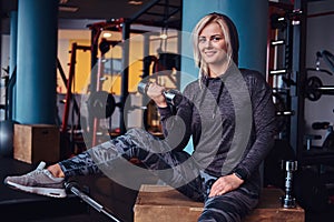 Sportive girl with blonde hair wearing hoodie sitting on a wooden box and holding a dumbbell, smiling and looking at a
