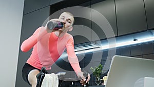 Sportive male athlete drinking water after hard training and intensively pedaling on stationary smart bicycle at home in the kitch