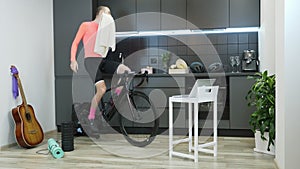Sportive man doing cycling sports exercises on indoor smart trainer and rubbing face with towel after hard training staying at hom