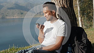 Sportive man tourist sitting on grass with backpack making audio message voice note ai photo on smart phone and enjoying