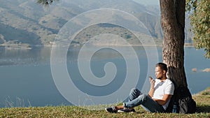 Sportive man tourist sitting on grass with backpack making audio message voice note ai photo on smart phone and enjoying
