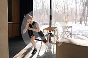 Sportive slim female doing single leg lunge using chair in living room. Side view of motivated fitness woman performing