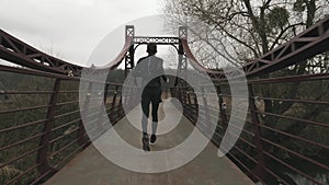 Sportive thin man runs on bridge across river in park, backside view. Male athlete running outdoor. Young guy in black tights and