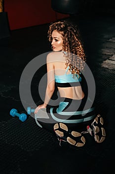 Sportive woman with blond curly hair in elegant sportive suit trains in gym with dumbbells