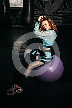 Sportive woman with blond curly hair in elegant sportive suit trains in gym with fitball