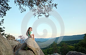 Sportive woman sitting alone taking a break on hillside trail. Female hiker enjoying view of evening nature from rocky