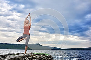 Sportive woman standing on the rock and practicing yoga against river.