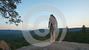 Sportive woman walking alone on hillside trail. Female hiker enjoying view of evening nature from rocky cliff on