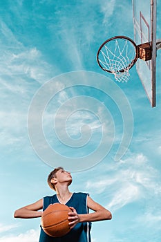Sports and basketball. A young teen in a black tracksuit playing basketball on the school Playground. Cloudy blue sky with basket