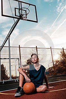 Sports and basketball. A young teenager in a black tracksuit poses with a basketball on a sports field while sitting on the ground