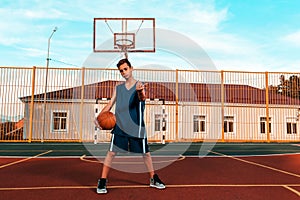Sports and basketball. A young teenager in a blue tracksuit confidently poses with a ball, standing on the Playground, and beckons