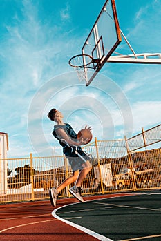 Sports and basketball. A young teenager in a blue tracksuit throws a ball into the basket in a powerful jump. Blue sky in the