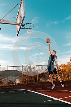 Sports and basketball. A young teenager in a blue tracksuit throws a jump ball into the basket. Blue sky. Vertical