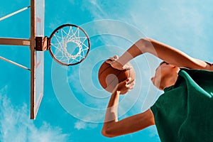 Sports and basketball. A young teenager in a green tracksuit throws a ball into the basket. Bottom view. Blue sky in the