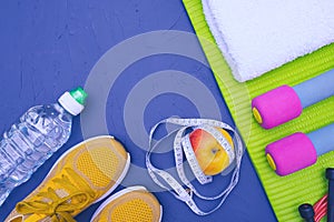 Sports equipment - green mat, bottle of water and towel, sneakers and dumbbells, apple and measurement tape, lie on blue, textured