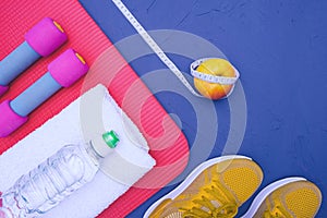 Sports equipment - mat, bottle of water and towel, sneakers, apple and measurement tape, lie on blue, textured background