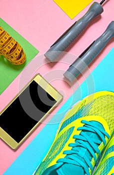 Sports equipment with running shoes, skipping rope, mobile phone with black screen and measuring tape on colorful background.