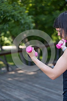 Sports girl holds small womens pink dumbbells. Young woman does outdoor fitness. Copy space. Vertical frame