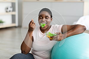 Sports and healthy eating for slimming. Plump black woman with fitness ball enjoying tasty vegetable salad at home