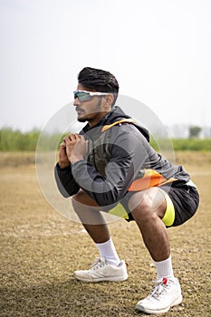 Sports and Healthy life concept - Indian young sports man with fitness clothing doing squats and exercising on a field