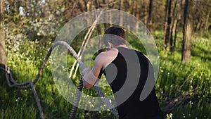 Sports man is training with undulating ropes. Hardwork. Black T shirt. Green park with trees. Sunnyday. Backside view