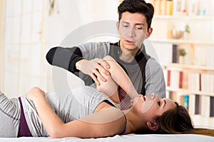 Sports massage. Hnadsome massage therapist massaging elbow of a female athlete, in a doctor office background
