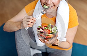 Sports and nutrition concept. Fit senior woman sitting on yoga mat, eating fresh vegetable salad after domestic workout