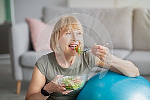 Sports and nutrition concept. Smiling senior lady leaning on fitness ball, eating fresh vegetable salad at home