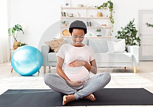 Sports and pregnancy concept. Lovely black expectant woman hugging her big belly while meditating on mat at home