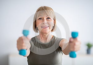 Sports during quarantine. Attractive senior woman doing fitness with dumbbells at home, exercising her arm muscles