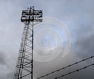 Sports stadium floodlights and barbed wire against stormy sky.