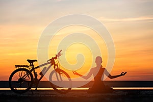 Sports woman doing yoga at sunrise on the sea beach against the background of orange sky and bicycle. Fitness concept. Lotus postu