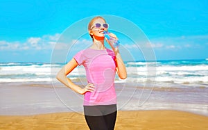 sports woman drinks water from bottle on the beach near the sea