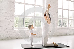 Sports young mother in white wear is engaged in fitness and yoga with a baby girl at light gym with big windows